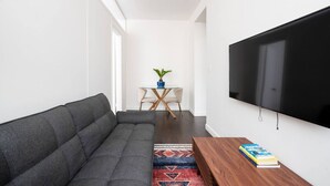 A modern living room with a gray sofa, a wooden coffee table with books, a TV mounted on the wall, and a small round table with chairs and a plant in the background. - 2 Bedroom Gem in Financial (New York)
