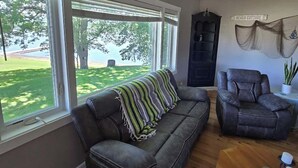 A living room with a leather sofa, a recliner, and a coffee table. There is a large window with a view of a lawn and trees. A sign on the wall reads "Beach Cottage."