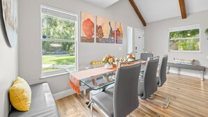 A dining area with a glass table, grey chairs, and a bench. There are three paintings on the wall, a bench with a cushion, and a window with a view of greenery.