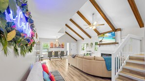 A spacious living room with a wooden ceiling, a large sofa, a staircase, and a neon sign.