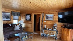 L’intérieur chaleureux d’un chalet avec un plafond en bois, un grand téléviseur à écran plat, un canapé, une table basse et une table à manger avec des chaises.