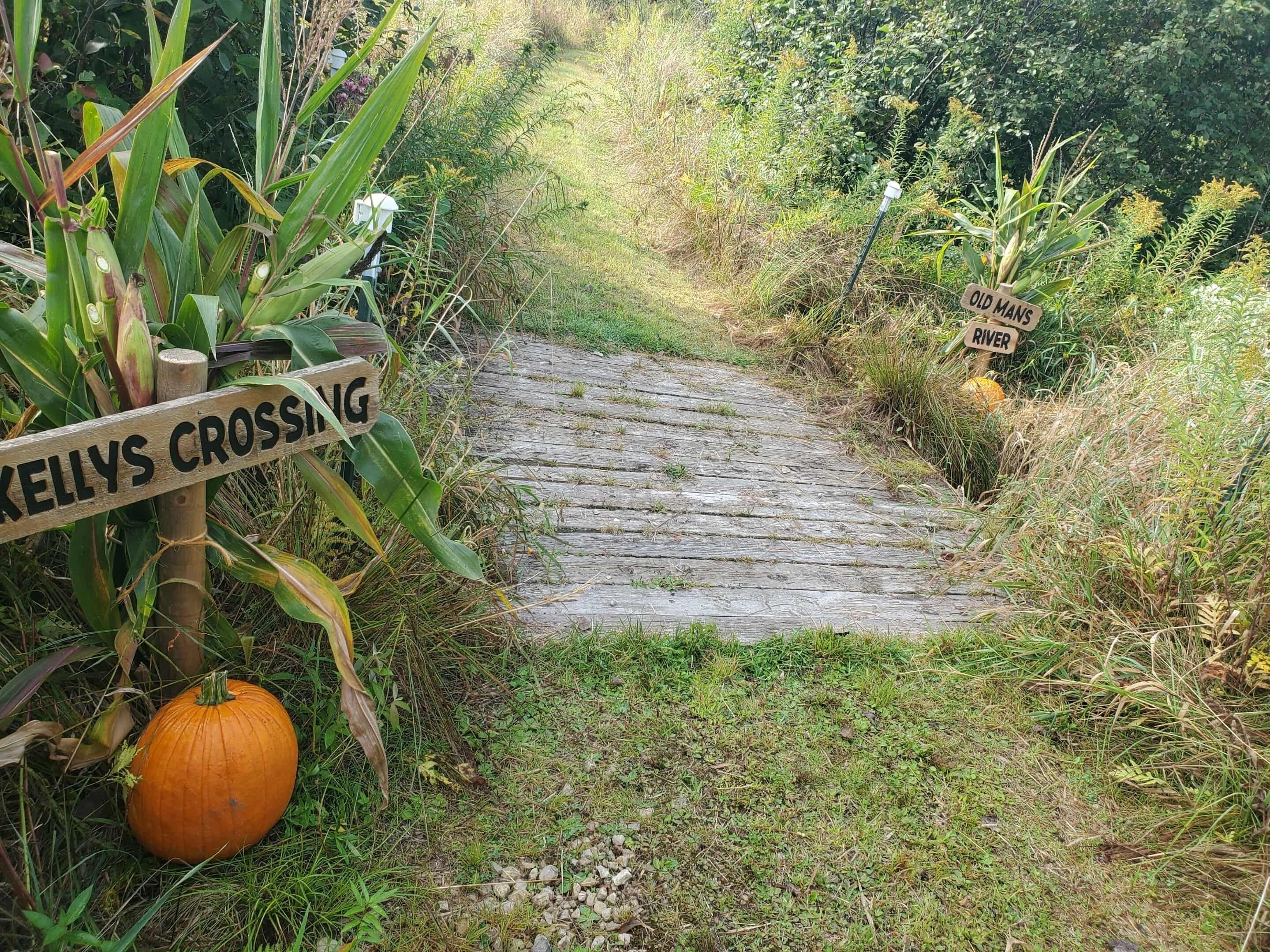 A rustic wooden bridge with signs indicating 'Kellys Crossing' and 'Old Mans River'.