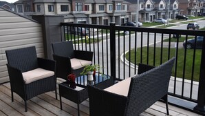 Un balcón con mobiliario de mimbre y una pequeña mesa, con vistas a una zona residencial con casas y coches aparcados.
