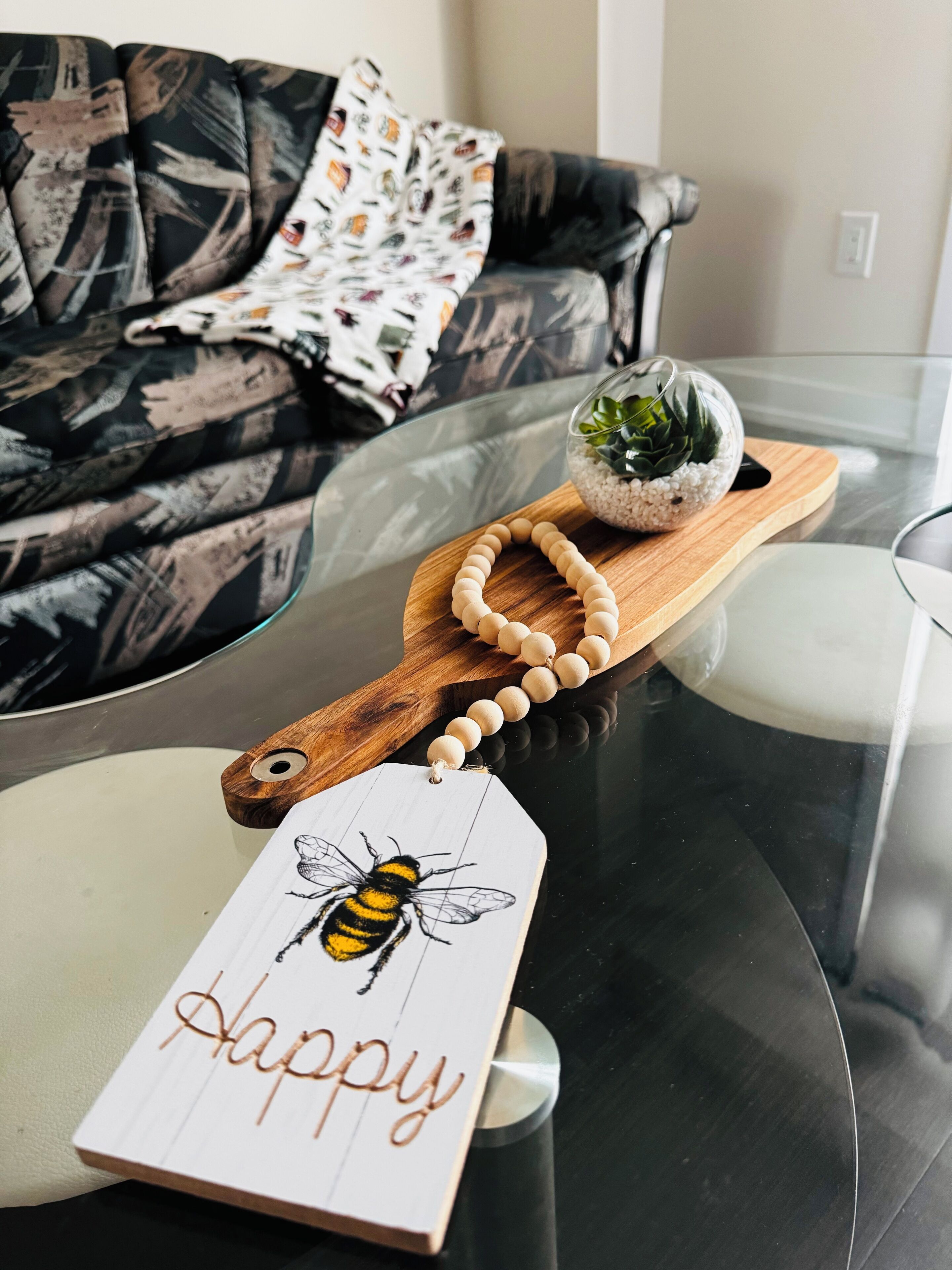 Une table en verre sur laquelle repose un plateau en bois, un collier de perles, un objet décoratif en verre contenant une plante, et une étiquette en bois ornée d’une illustration d’abeille et du mot « heureux ».
