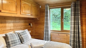 A wooden cabin room with a bed, plaid curtains, and a window overlooking greenery. - 3 bedroom accommodation in Rowardennan (Rowardennan)