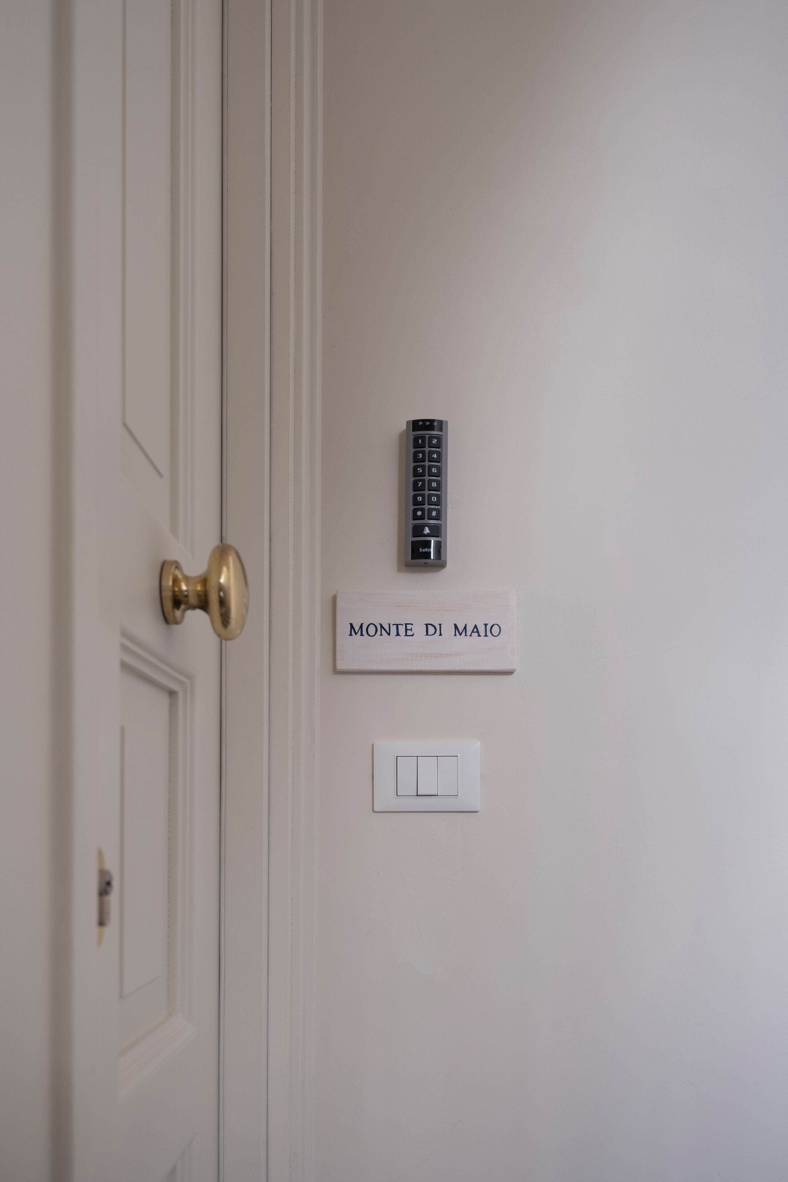 A white door with a brass handle, a wall-mounted intercom, and a light switch below a sign that reads "MONTE DI MAIO".