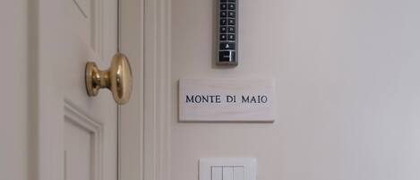 A white door with a brass handle, a wall-mounted intercom, and a light switch below a sign that reads "MONTE DI MAIO".