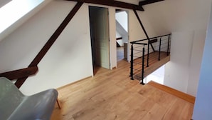 A loft-style room with a sloped ceiling, wooden beams, and a mezzanine with a black metal railing. - Gîte des trois châteaux (Husseren-les-Châteaux)
