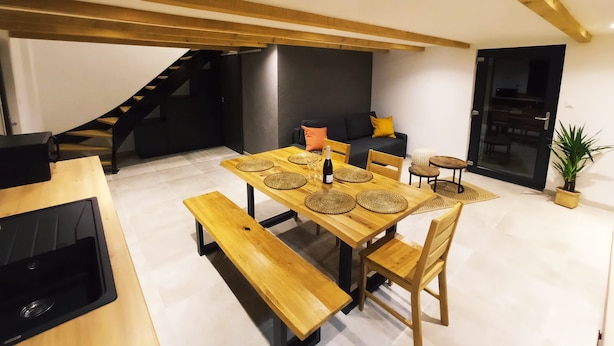 A modern dining area with a wooden table, benches, and a black countertop. - Gîte des trois châteaux (Husseren-les-Châteaux)
