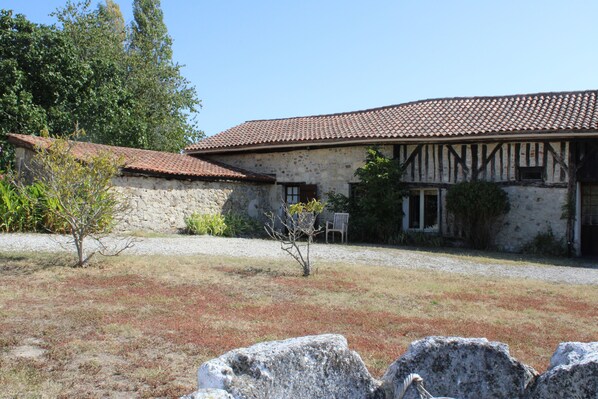 A stone building with a tiled roof, a gravel driveway, and a small tree in the foreground.
