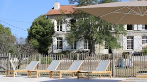 A pool area with lounge chairs and an umbrella, surrounded by a wooden fence and a stone building with a tiled roof. - Gîtes au Château de Servanches, Dordogne, 20 Personnes, Piscine & Nature (Servanches)