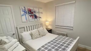 Un dormitorio con una cama, una mesita de noche, un radiador, una ventana con persianas y dos cuadros abstractos en la pared.