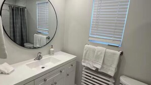 Un baño moderno con un tocador blanco, un espejo redondo, un inodoro y una ventana con persianas.