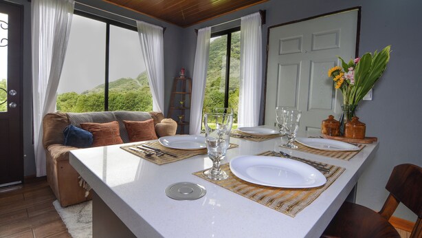 Un comedor con una mesa dispuesta para cuatro personas, un sofá con cojines y vistas a zonas verdes a través de las ventanas.
