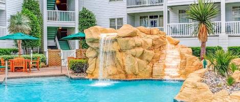A pool with a waterfall feature, surrounded by a white building with balconies and outdoor seating.