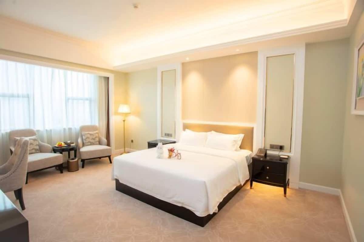 Habitación de hotel con una cama grande, dos sillones, una mesita de noche, una lámpara y una ventana con cortinas.