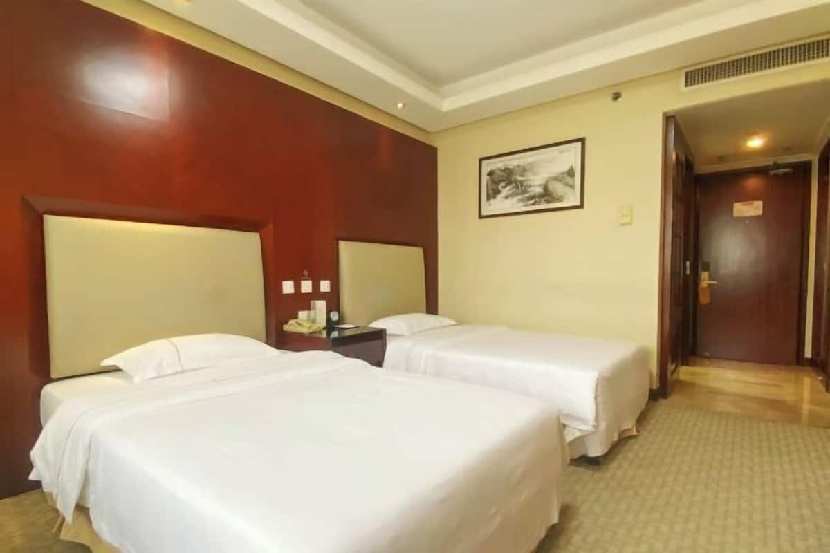 Habitación de hotel con dos camas, cabecera de madera, mesita de noche con lámpara, un cuadro enmarcado en la pared y una puerta que da a otra habitación.