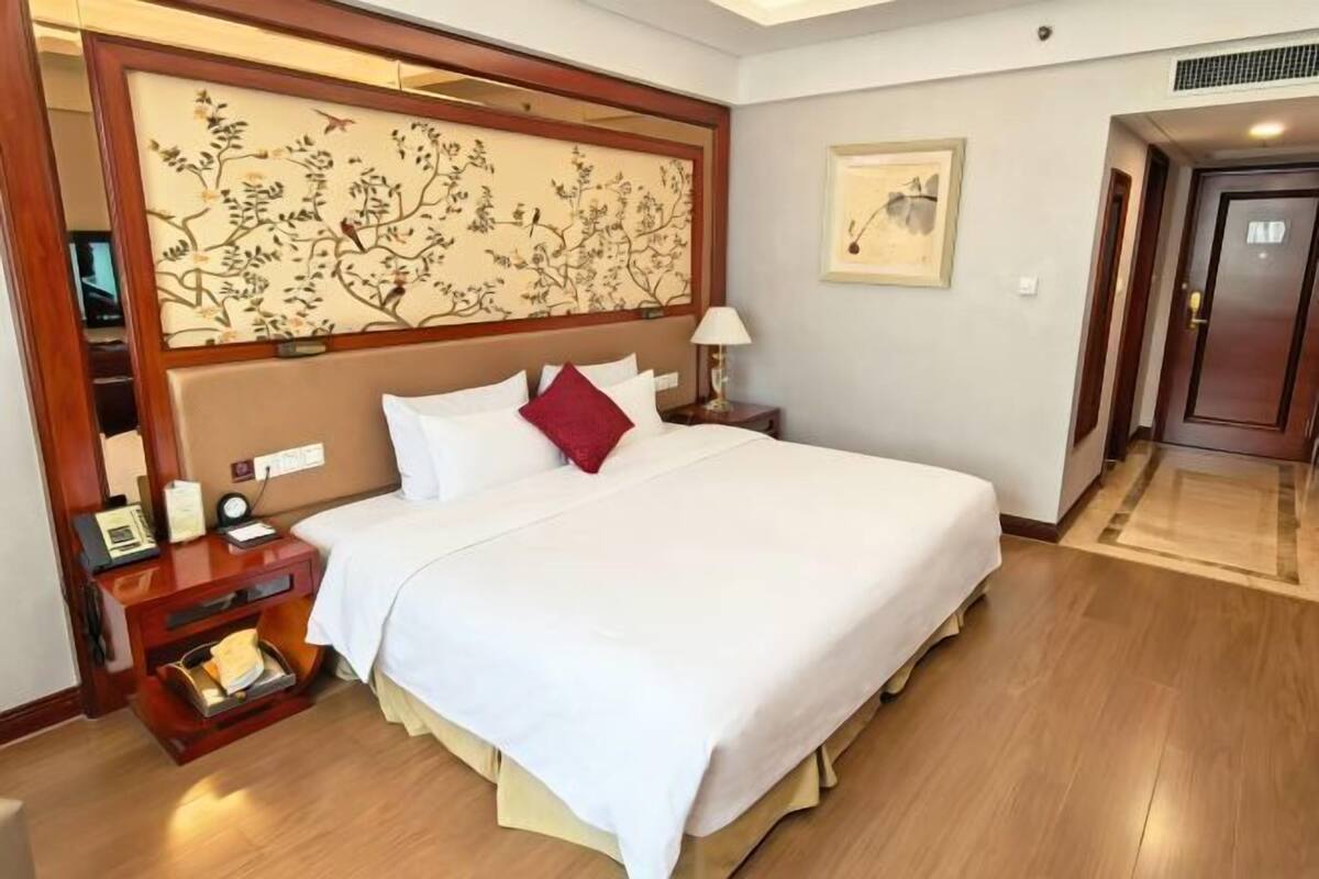 Una habitación de hotel con una cama grande, una mesita de noche con lámpara, una obra de arte en la pared y una puerta.