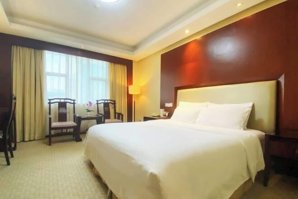 Habitación de hotel con una cama grande, un escritorio con una silla, una lámpara y un ventanal con cortinas.