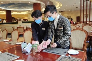 Deux personnes sont assises à une table dans un restaurant d’hôtel au décor moderne, avec un espace de réception visible à l’arrière-plan.