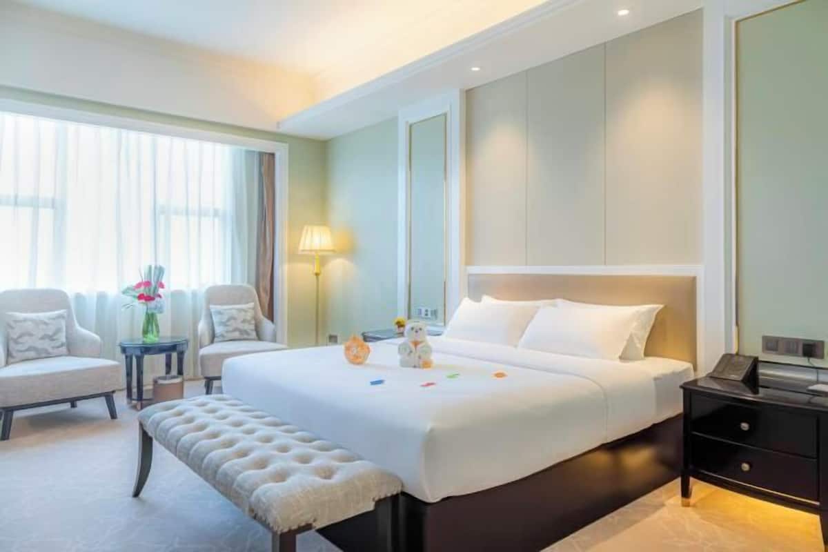 Una habitación de hotel con una cama grande, dos sillones, una mesita pequeña, un banco y una mesita de noche.