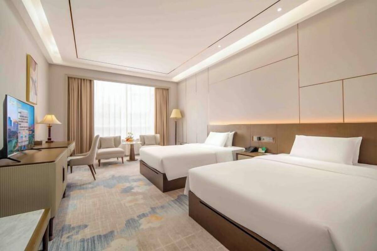 Habitación de hotel con dos camas, un escritorio con ordenador, una mesita con lámpara y vistas a la ciudad a través de amplias ventanas.