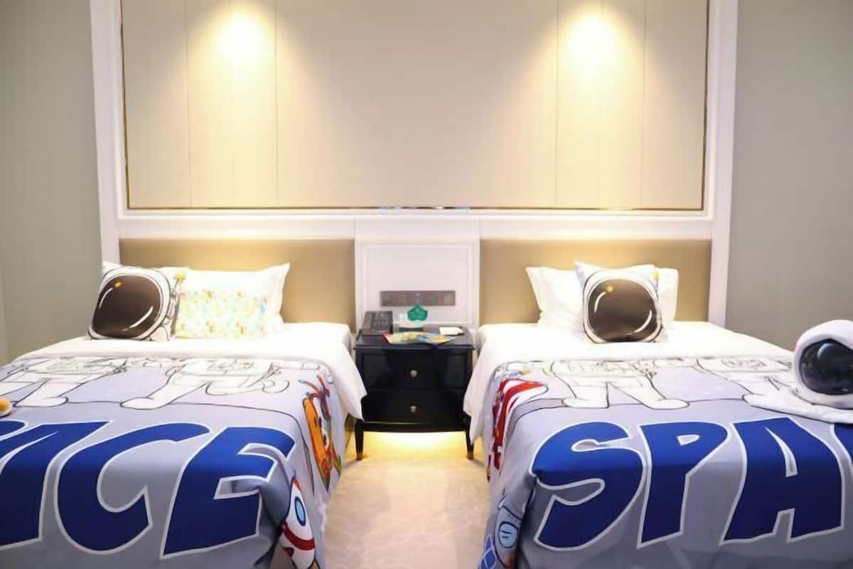Habitación de hotel con dos camas, cada una con una colcha grande con motivos de 'SPA' e 'ICE'. Hay una mesita de noche entre las camas, con una lámpara y una pequeña mesa.