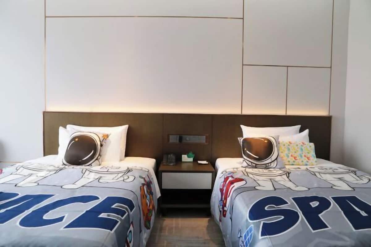 Habitación de hotel con dos camas, cada una con una colcha decorativa, un cabecero de madera, una mesita de noche y una pequeña mesa.