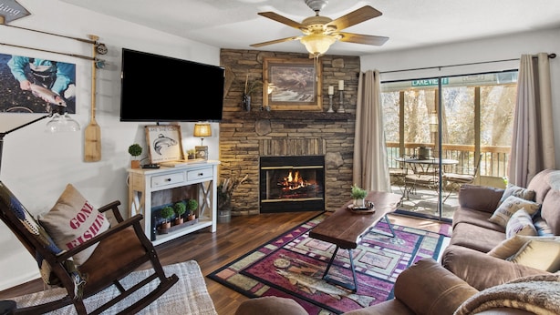Una sala de estar con chimenea de piedra, ventilador de techo, televisor y vistas al lago.