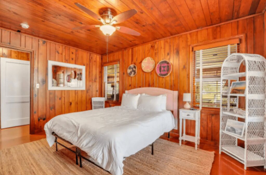A wooden-paneled bedroom with a bed, a white bedside table, a ceiling fan, and a white shelving unit.