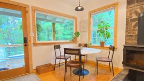 A cozy dining area with a round table and four chairs, large windows with a view of greenery, a stone fireplace, and a wooden ceiling. - 3 Miles to Asheville! Sunfilled Cabin with Wi/Fi (Asheville)