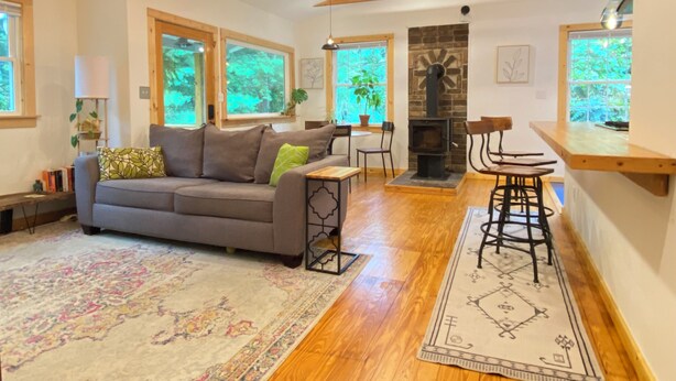A cozy living room with a sofa, a coffee table, and a fireplace. There are wooden beams on the ceiling and large windows with views of greenery. - 3 Miles to Asheville! Sunfilled Cabin with Wi/Fi (Asheville)