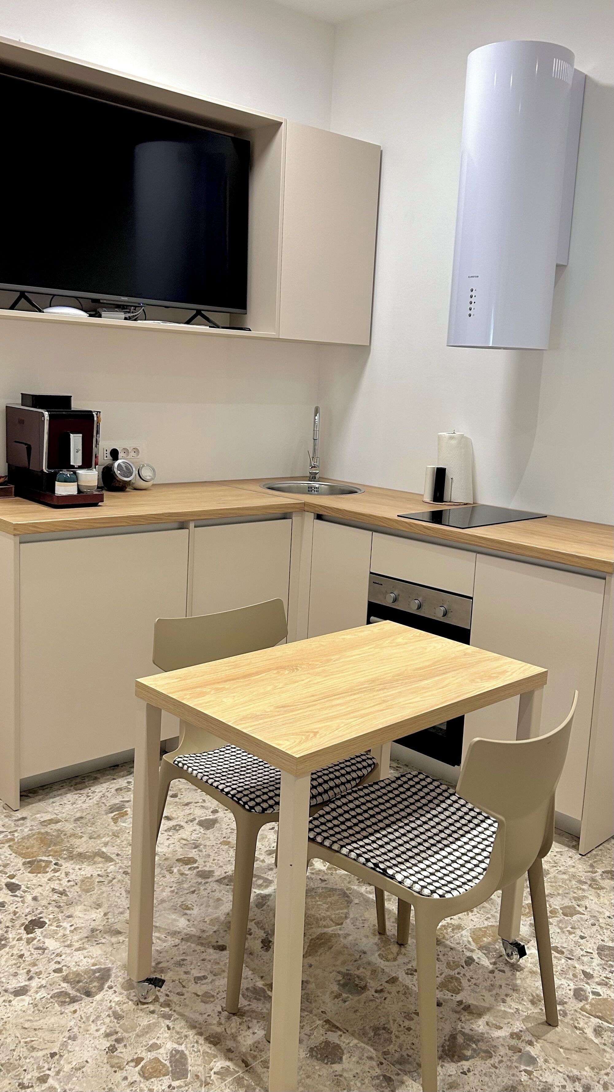 A compact kitchen with a wooden table and chairs, a built-in oven, and a large flat-screen TV above the counter.