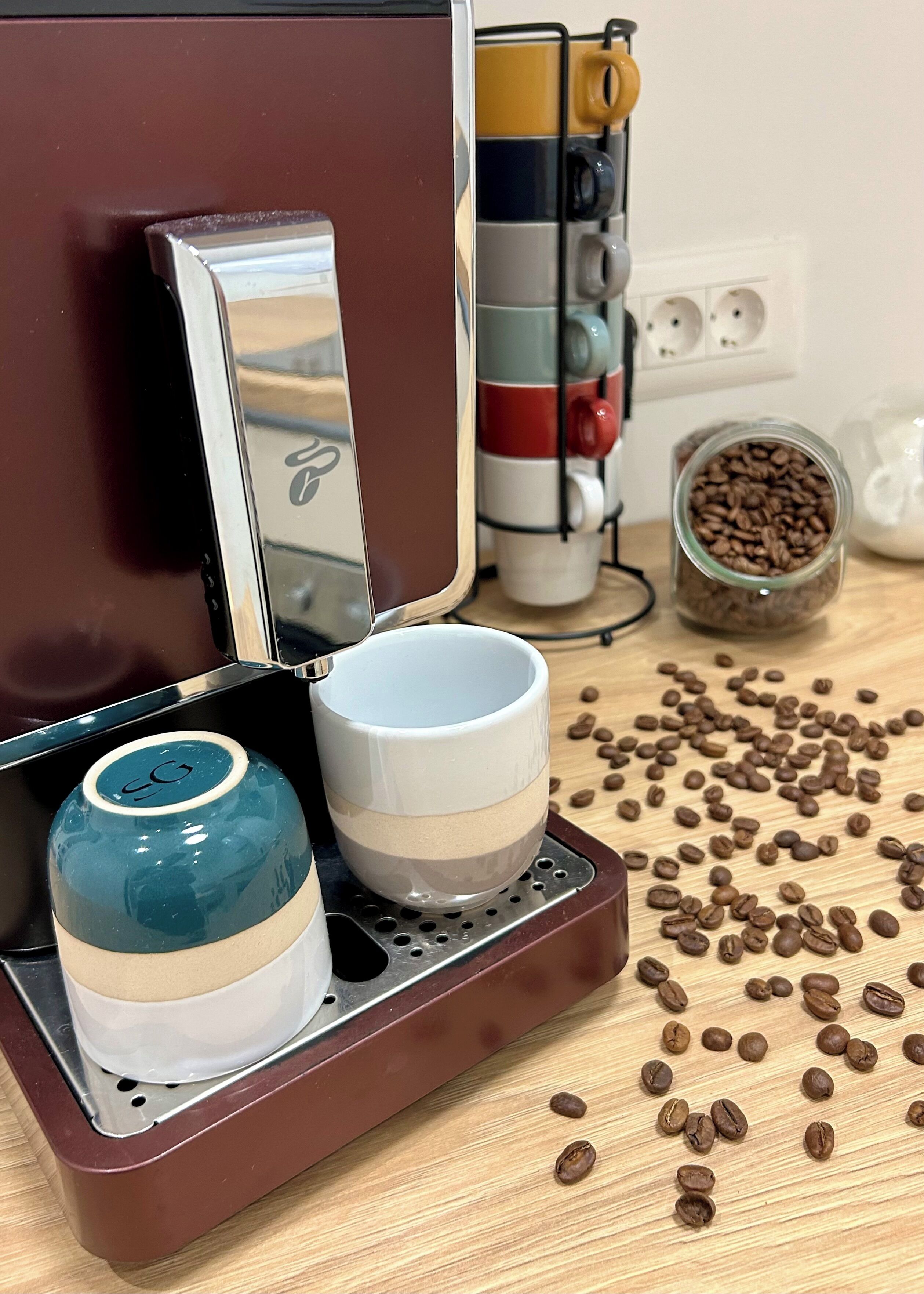 En kaffemaskine med vandbeholder og kaffekande, en kop samt en beholder med kaffebønner på en træoverflade.
