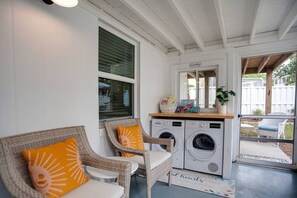 A laundry area with two washing machines, wicker chairs with cushions, a small table, and a window with blinds.