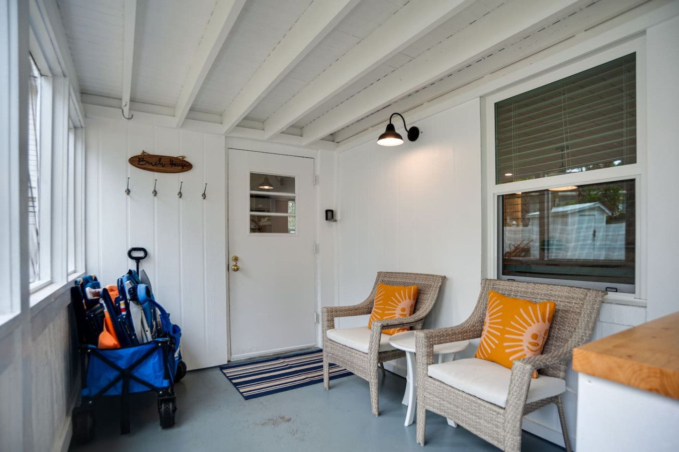 A room with a white door, two wicker chairs with orange cushions, a blue stroller, and a window with blinds.