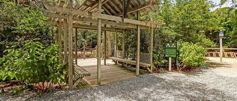 Eine Pergola aus Holz mit Bänken, umgeben von Kies und Pflanzen.
