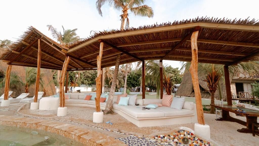 A covered outdoor seating area with a pool, palm trees, and a thatched roof structure.