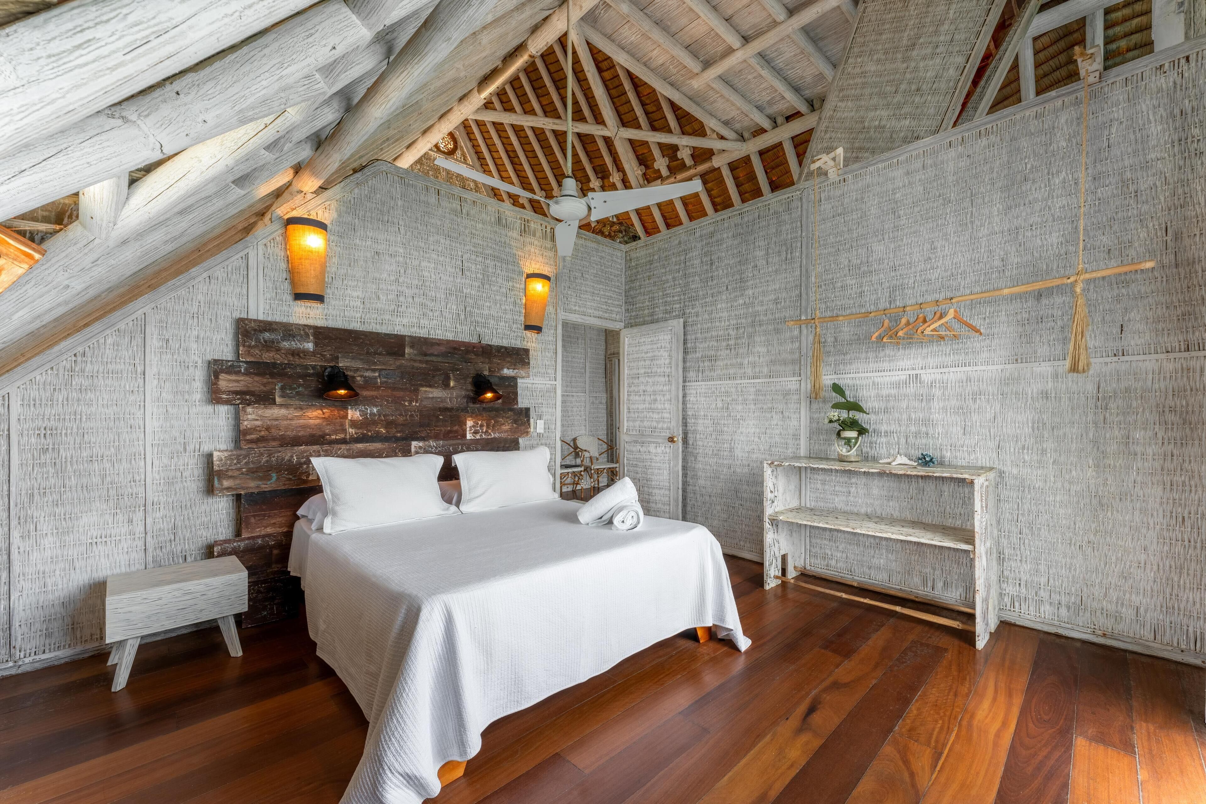 Un dormitorio con techo inclinado, vigas de madera, una cama con ropa de cama blanca, una mesita de noche pequeña y un banco.