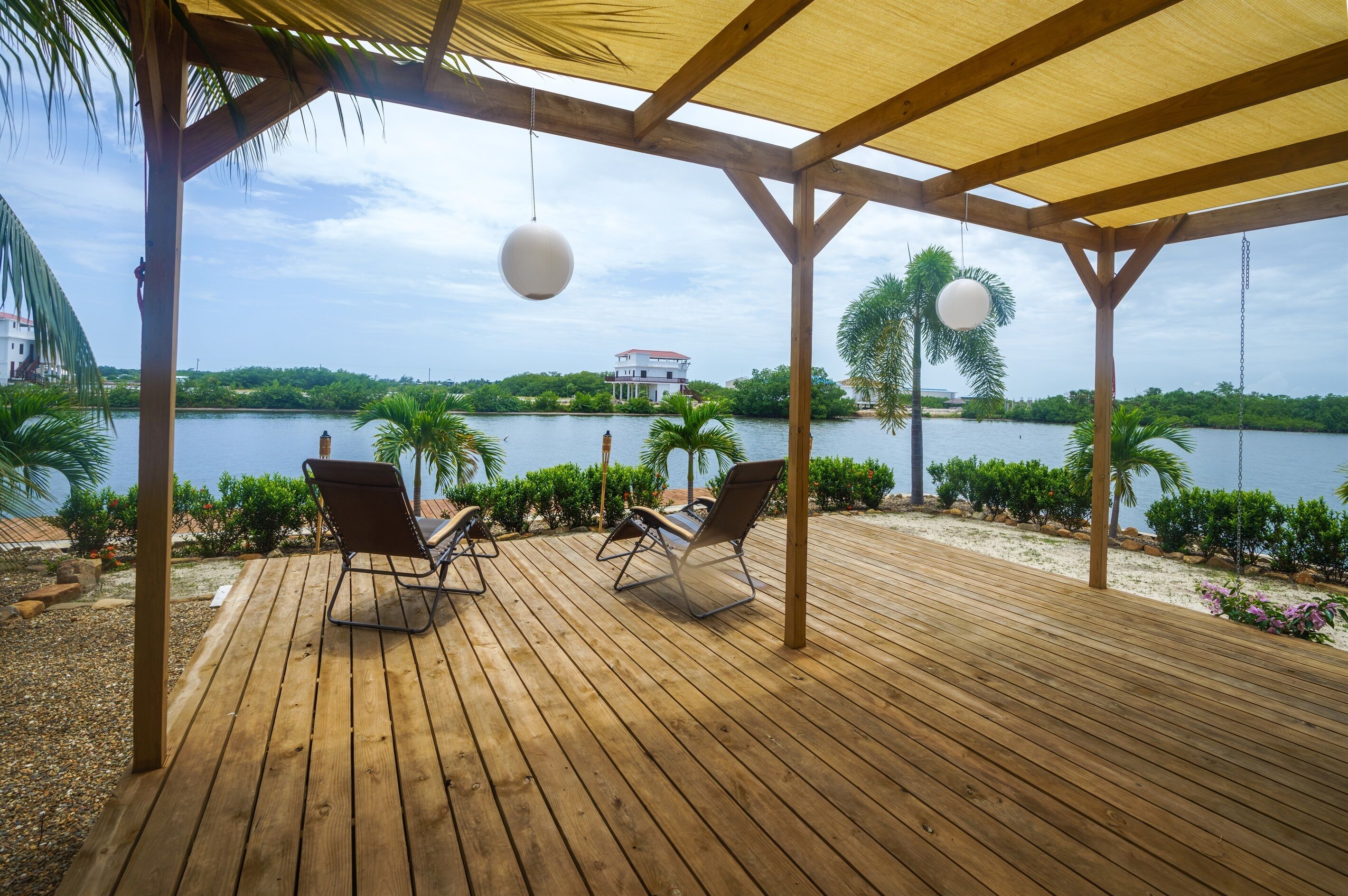 A wooden deck with lounge chairs overlooking a lake, surrounded by greenery and a white building in the distance.