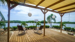 Een houten terras met lounge stoelen met uitzicht op een meer, omgeven door groen en een wit gebouw in de verte.
