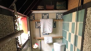 A narrow bathroom with a toilet, a sink, and a shower area. There is a towel hanging on a rack, and a shelf above the sink holds various items.