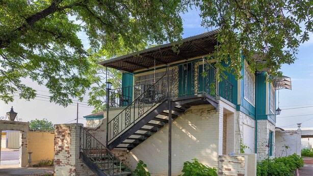A two-story building with a staircase leading to a balcony, surrounded by greenery and a brick wall. - Mariposa Monarch Walk to River (Kerrville)