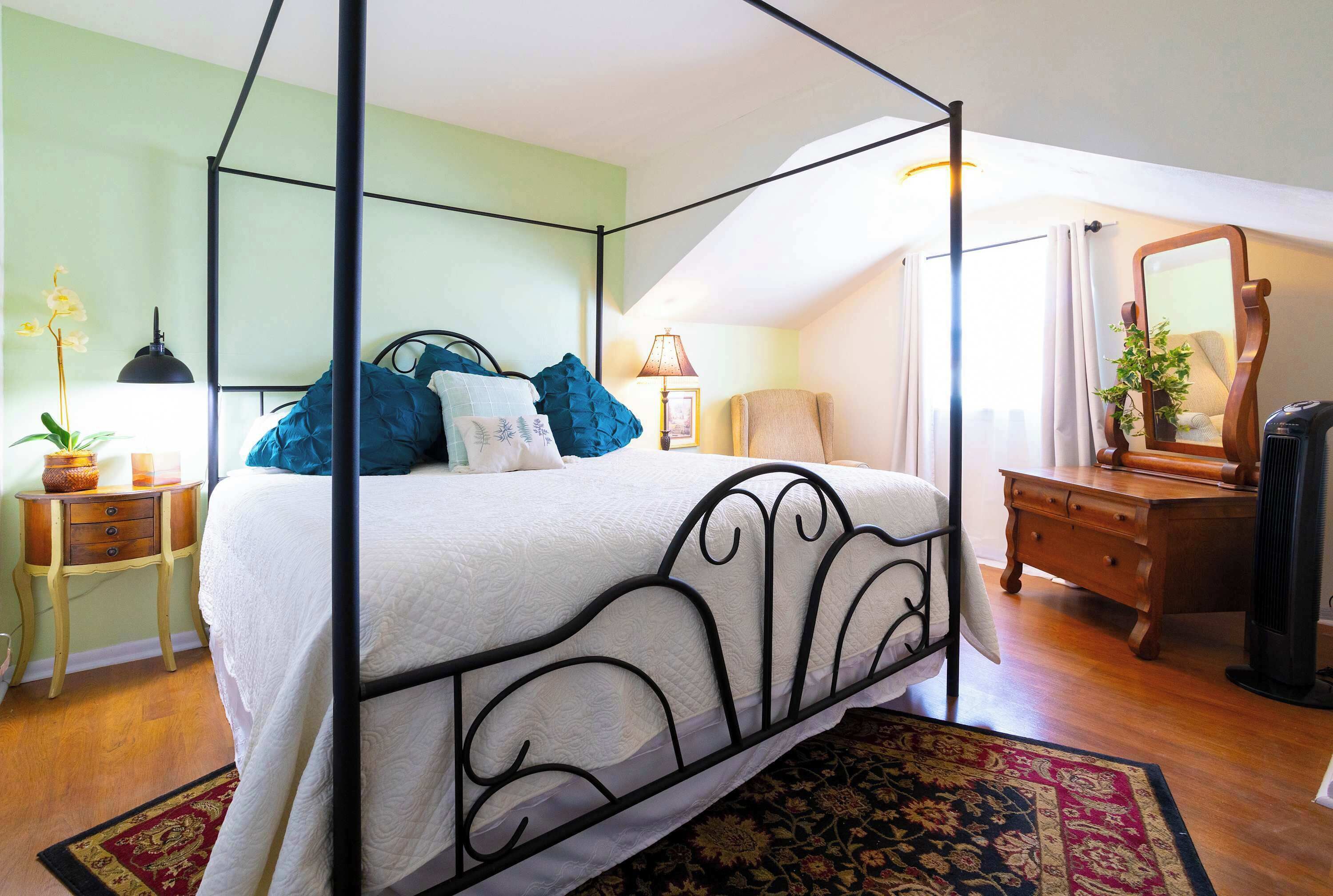 A four-poster bed with a white bedspread, a wooden nightstand with a lamp, a dresser, and a mirror.