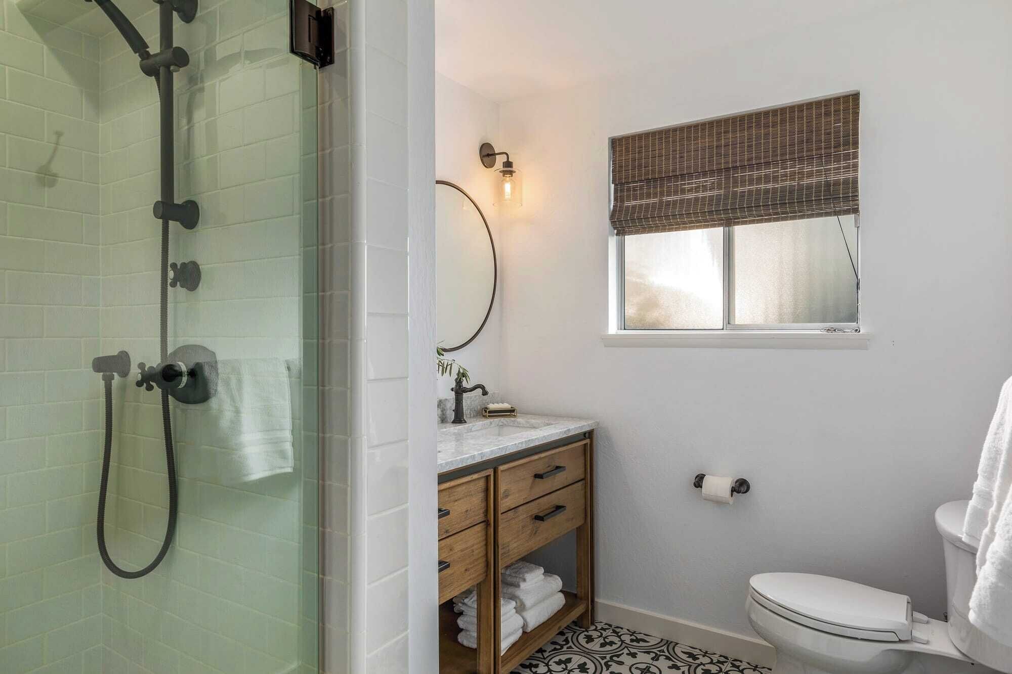 A bathroom with a glass shower enclosure, a wooden vanity with a mirror, a toilet, and a window with a bamboo blind.