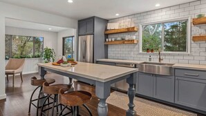 Moderni keittiö, jossa on keskeinen saareke, harmaat kaapit ja suuri ikkuna, josta avautuu näkymä puita kohti.