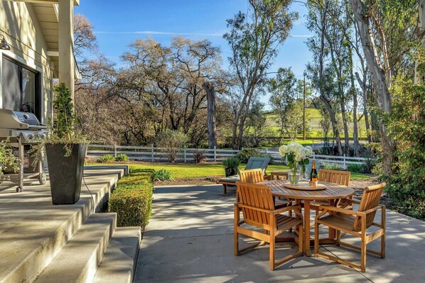 Un patio al aire libre con una mesa y sillas de madera, una parrilla y una valla blanca al fondo.