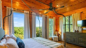 Een houten slaapkamer met een bed, gordijnen, een televisie en uitzicht op het bos.