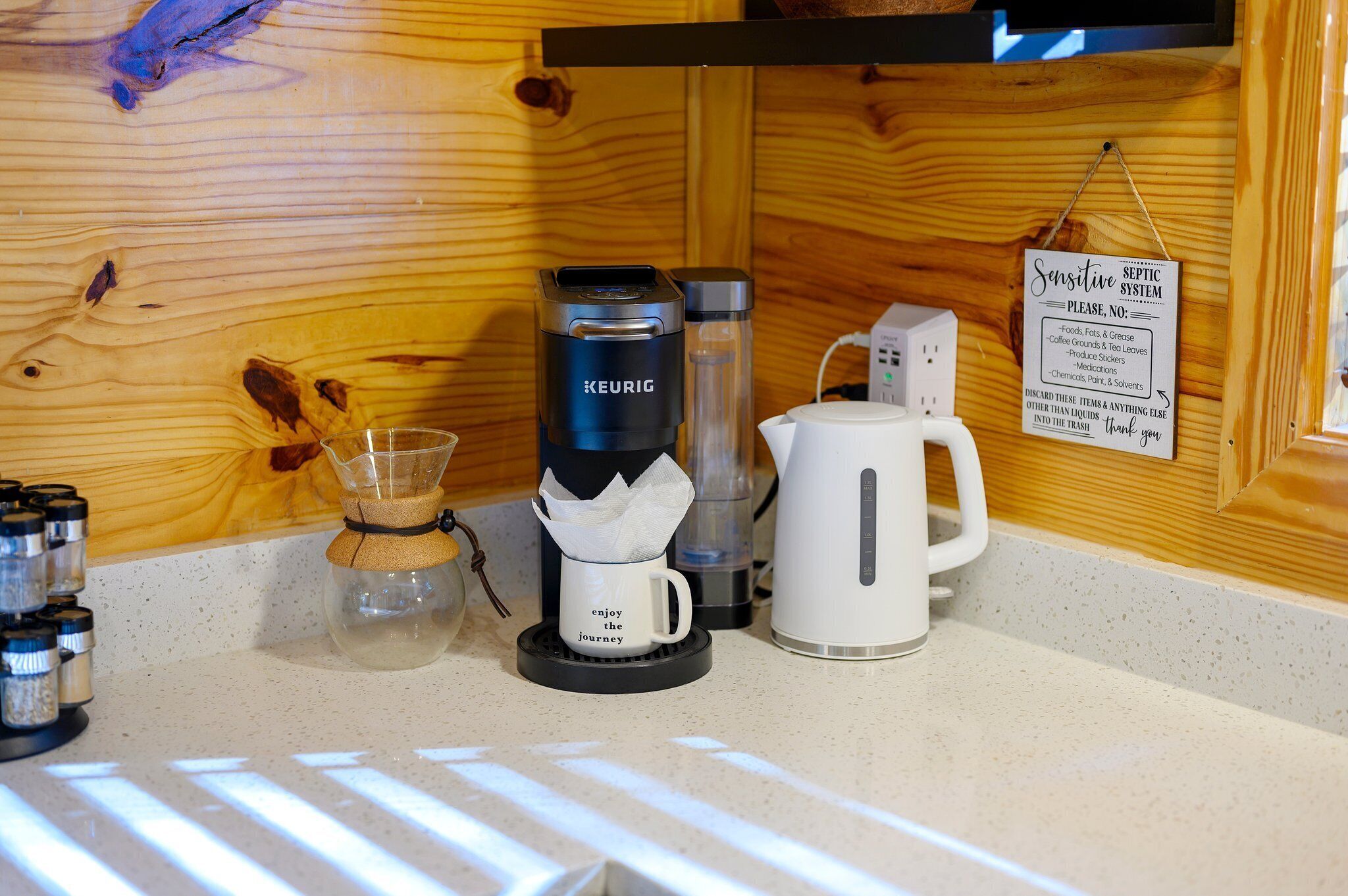 Et tekøkken med en Keurig kaffemaskine, en glaskande, en hvid kaffepotte og et skilt, der angiver et skrøbeligt septisk system.