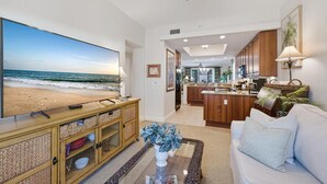 A spacious living room with a large flat-screen TV, a wooden entertainment unit, a white sofa, and a kitchen area with wooden cabinets and a central island. - Oversized 2BR Condo @ Resort near Beach (Naples)
